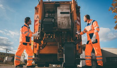 Zwei Männer in orangefarbener Arbeitskleidung bedienen ein großes orangefarbenes Müllfahrzeug im Freien.