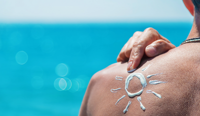 Eine sonnenförmige Zeichnung aus Sonnencreme auf einer nackten Schulter vor einem verschwommenen, blauen Meeres- und Himmelshintergrund.