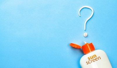 Eine geöffnete Sonnenschutzflasche mit orangefarbenem Deckel auf blauem Hintergrund, daneben eine aus Lotion geformte Fragezeichenform.