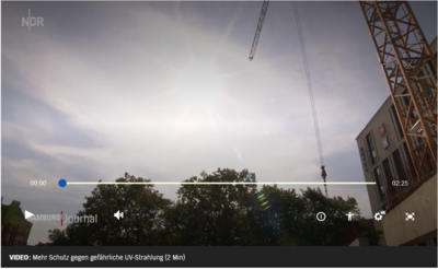 Ein Kran ragt in den Himmel, während die Sonne durch leichte Wolken scheint; Bäume und Gebäude sind im Vordergrund sichtbar.