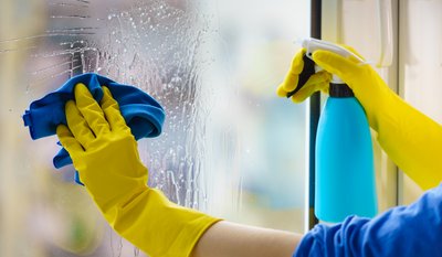Jemand mit gelben Gummihandschuhen reinigt ein Fenster mit einem blauen Lappen und einer Sprühflasche mit Reinigungsmittel.