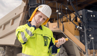 Ein Bauarbeiter in gelber Schutzkleidung und weißem Helm steht vor einem großen Fahrzeug und hält einen orangen Gegenstand, während er seinen Nacken berührt.